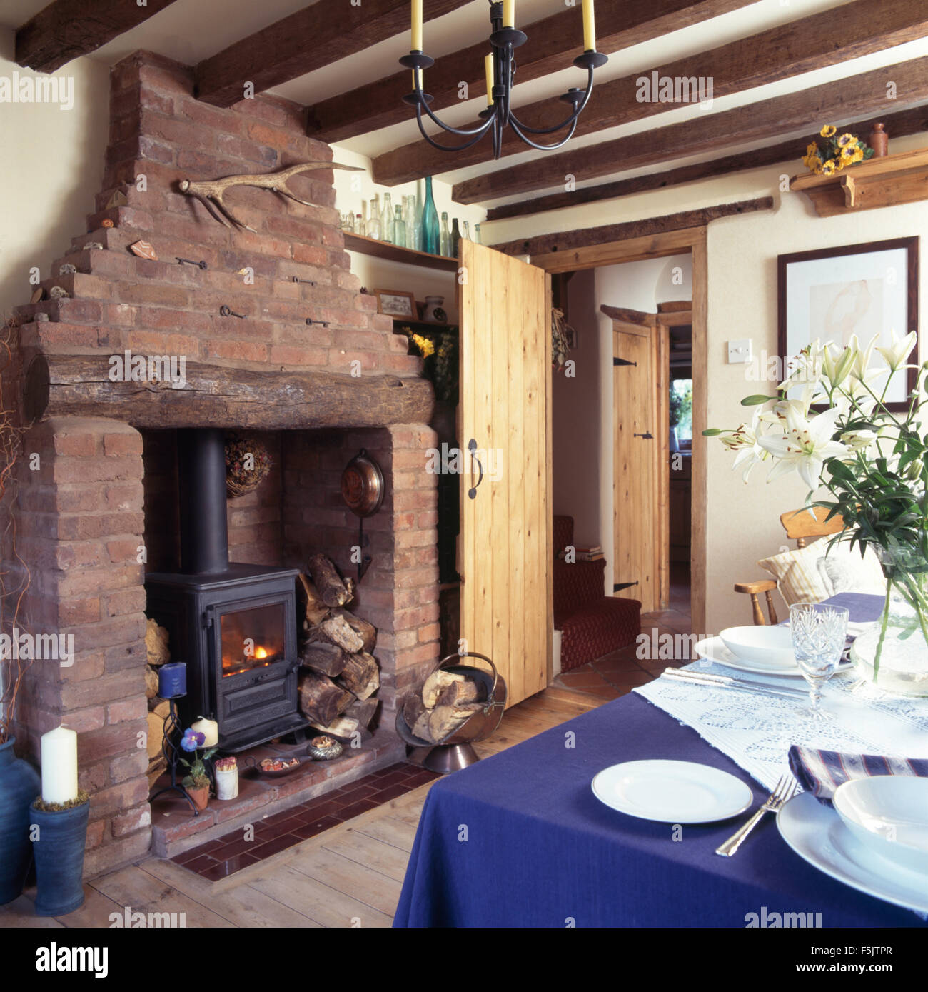 Wood burning stove in exposed brick fireplace in a cottage dining room with a blue cloth on table set for lunch Stock Photo