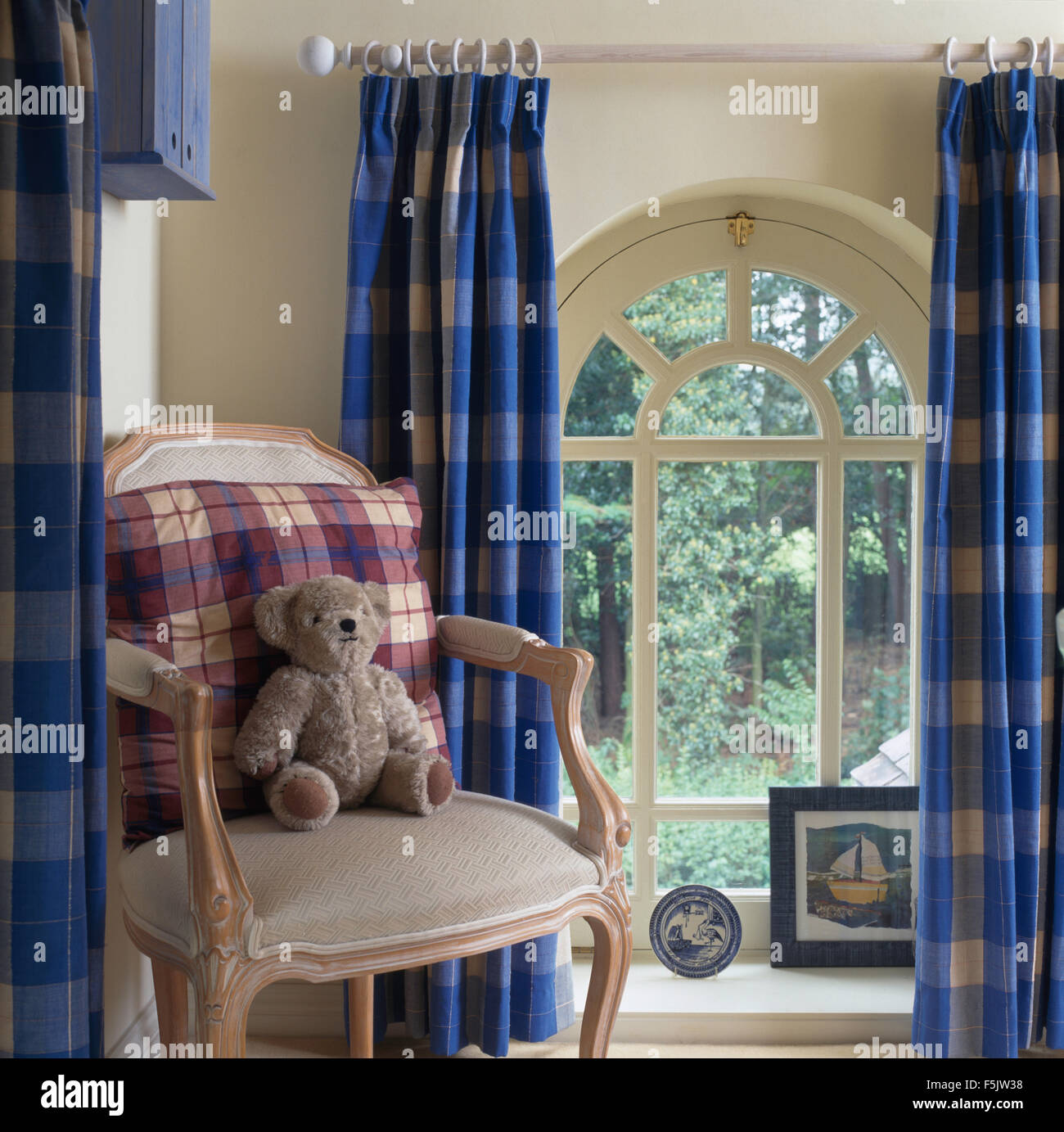 Teddy bear and checked cushion on chair beside an arched window with blue checked curtains in a country hall Stock Photo