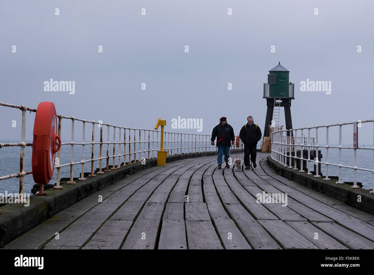 Couple walking pet dog on Whitby's West Pier, England, UK - grey tones of wooden decking boards, sea & sky in cold windy dull autumn weather at coast. Stock Photo