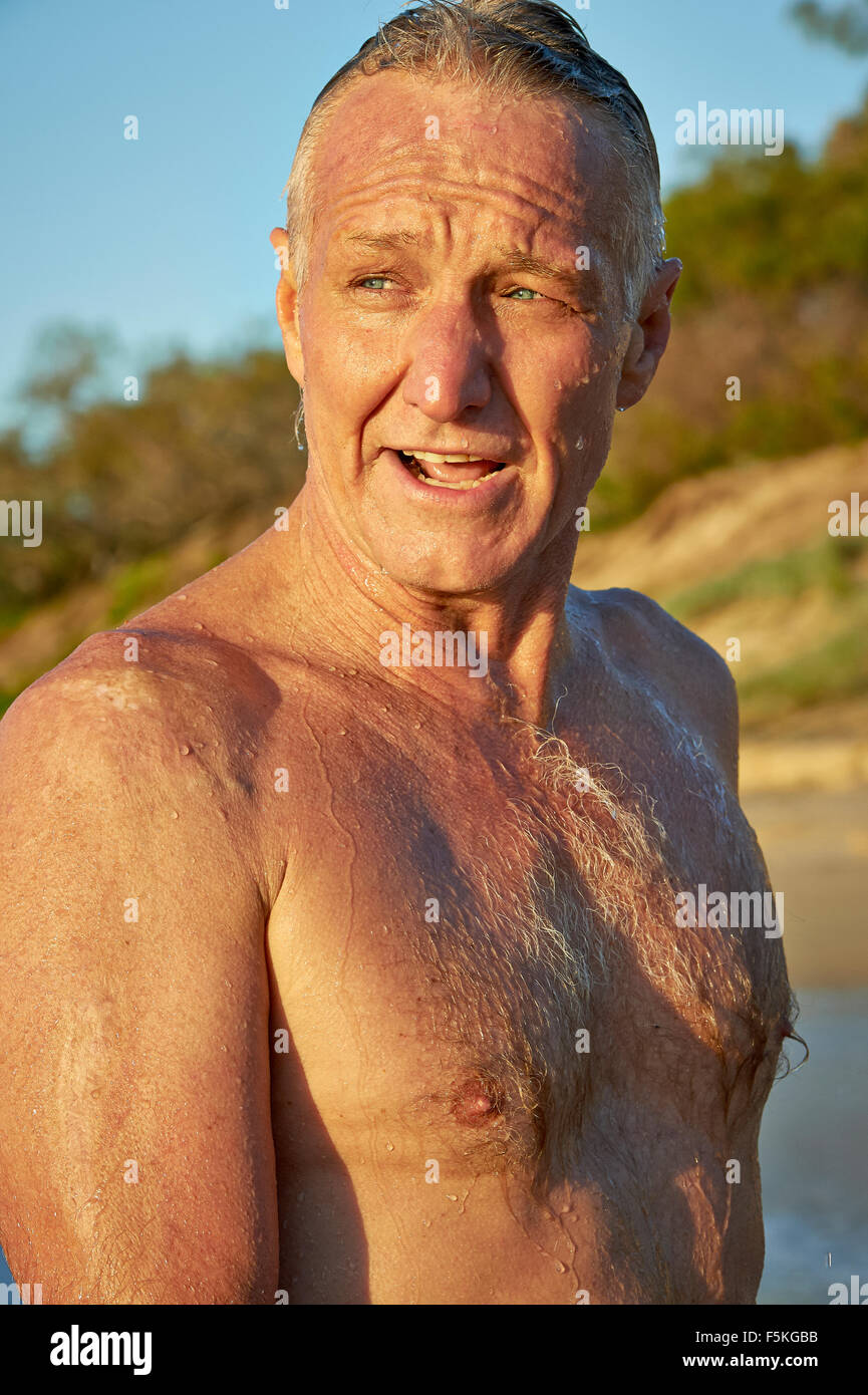 Mature aged man with blue eyes, walking on the beach just after a refreshing swim, water droplets all over him Stock Photo
