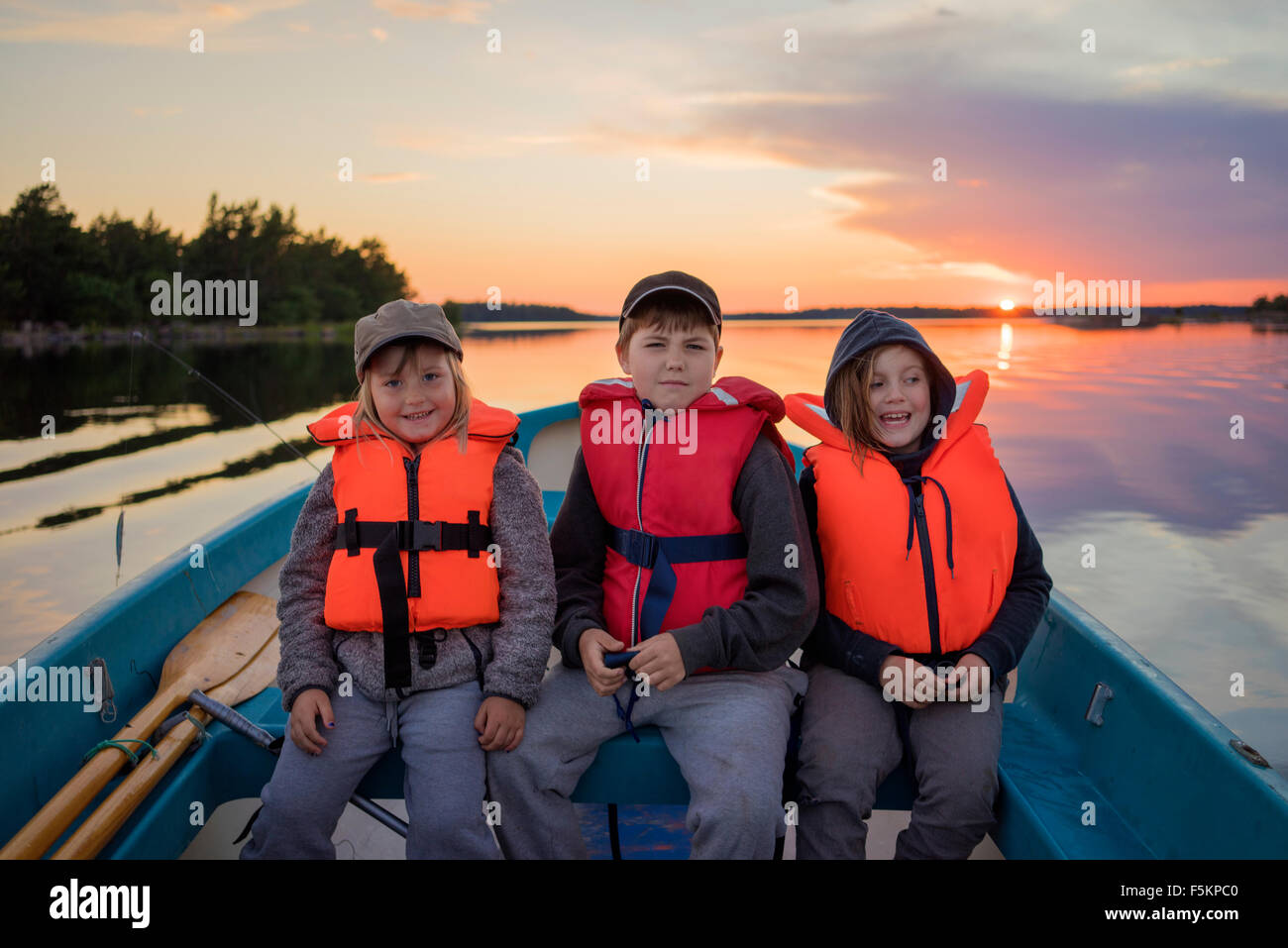 Children (4-5, 8-9, 10-11) wearing life jackets sitting in rowboat at sunset. Sweden Stock Photo
