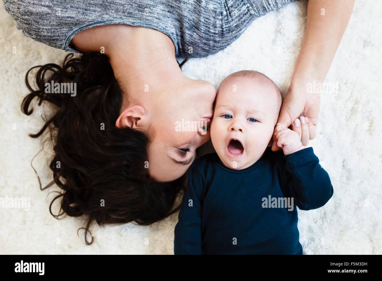 Mother lying on rug and kissing baby son on cheek, overhead view Stock Photo