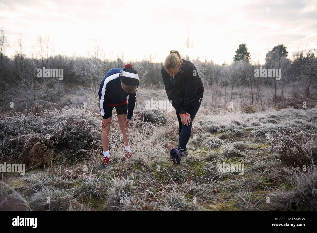 Mother and son on frosty grassland, hands on knees stretching legs looking down Stock Photo
