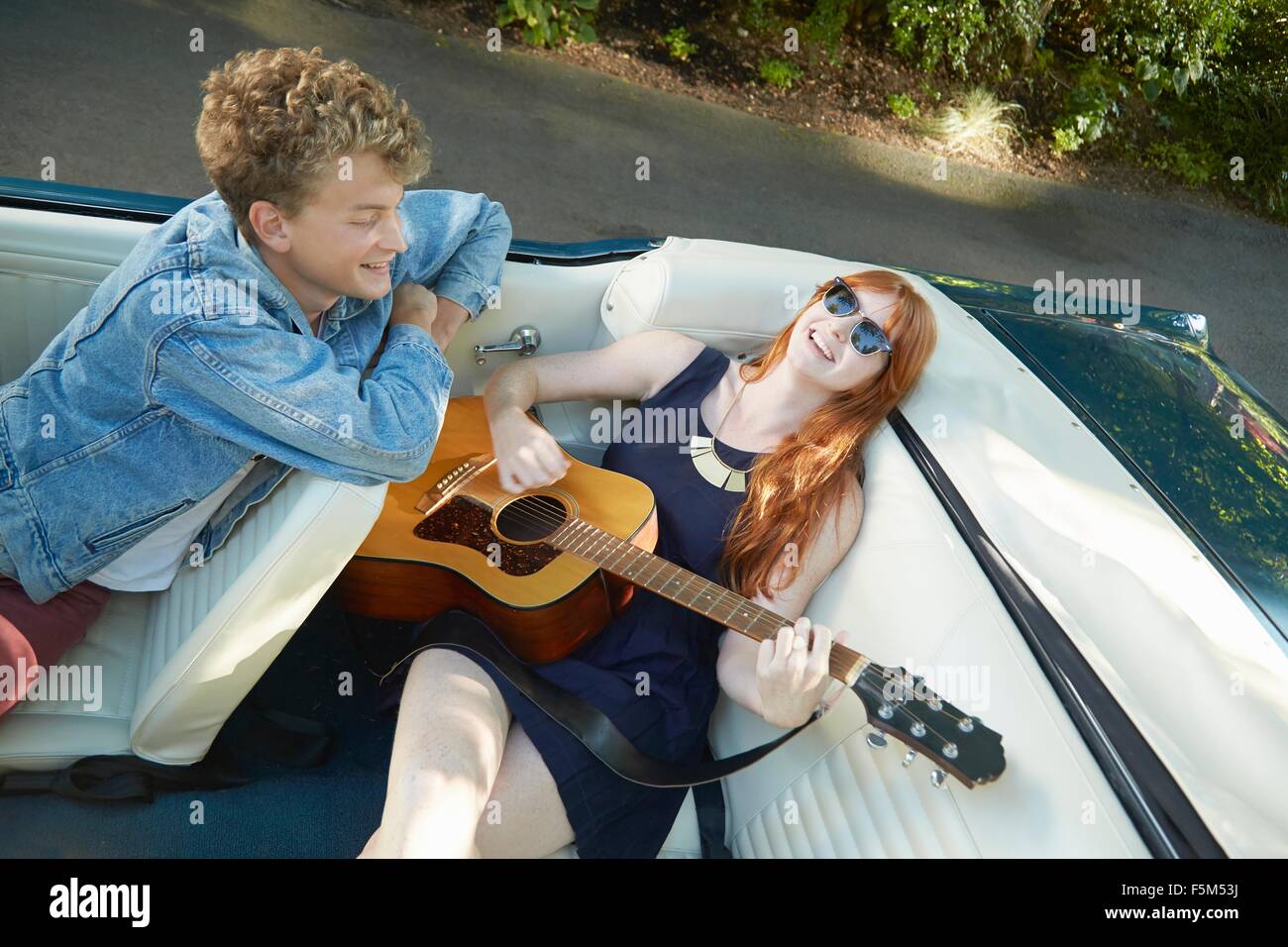 Young woman playing guitar for boyfriend in back seat of vintage convertible Stock Photo