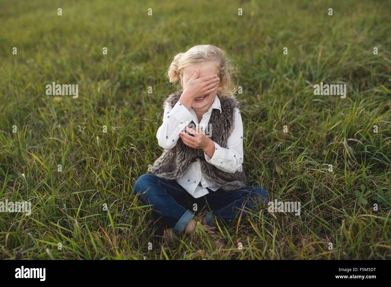 Young girl sitting on grass, covering eyes Stock Photo