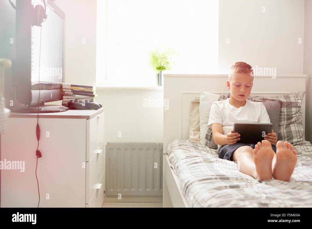 Boy sitting on bed looking down at digital tablet Stock Photo