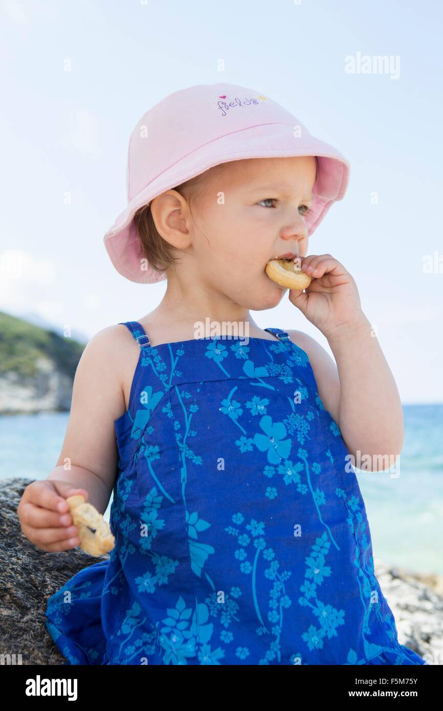 Female toddler wearing sunhat eating doughnut on beach Stock Photo