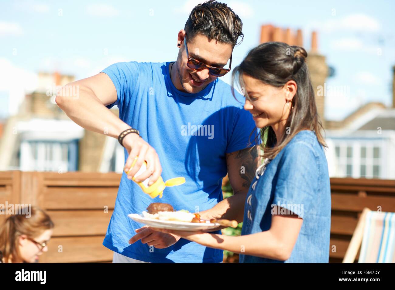 Mid adult man squirting mustard on burger at rooftop party Stock Photo