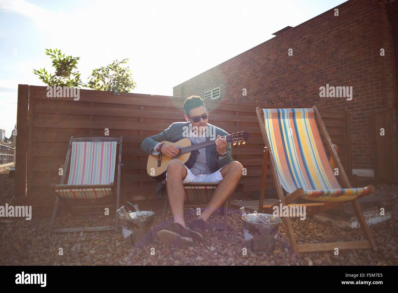 Mid adult man playing guitar on deckchair at rooftop party Stock Photo