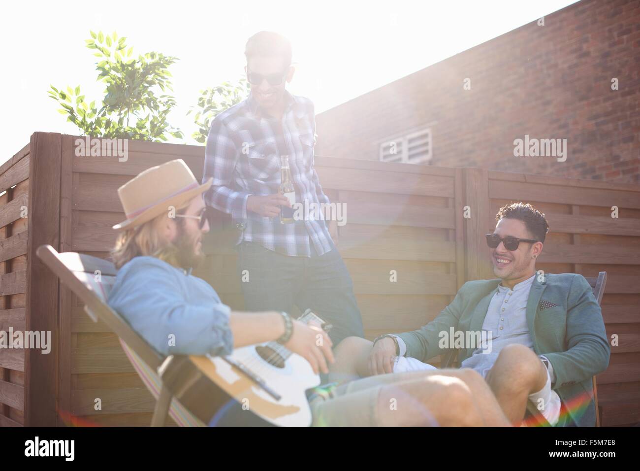 Three male friends chatting and playing guitar at rooftop party Stock Photo