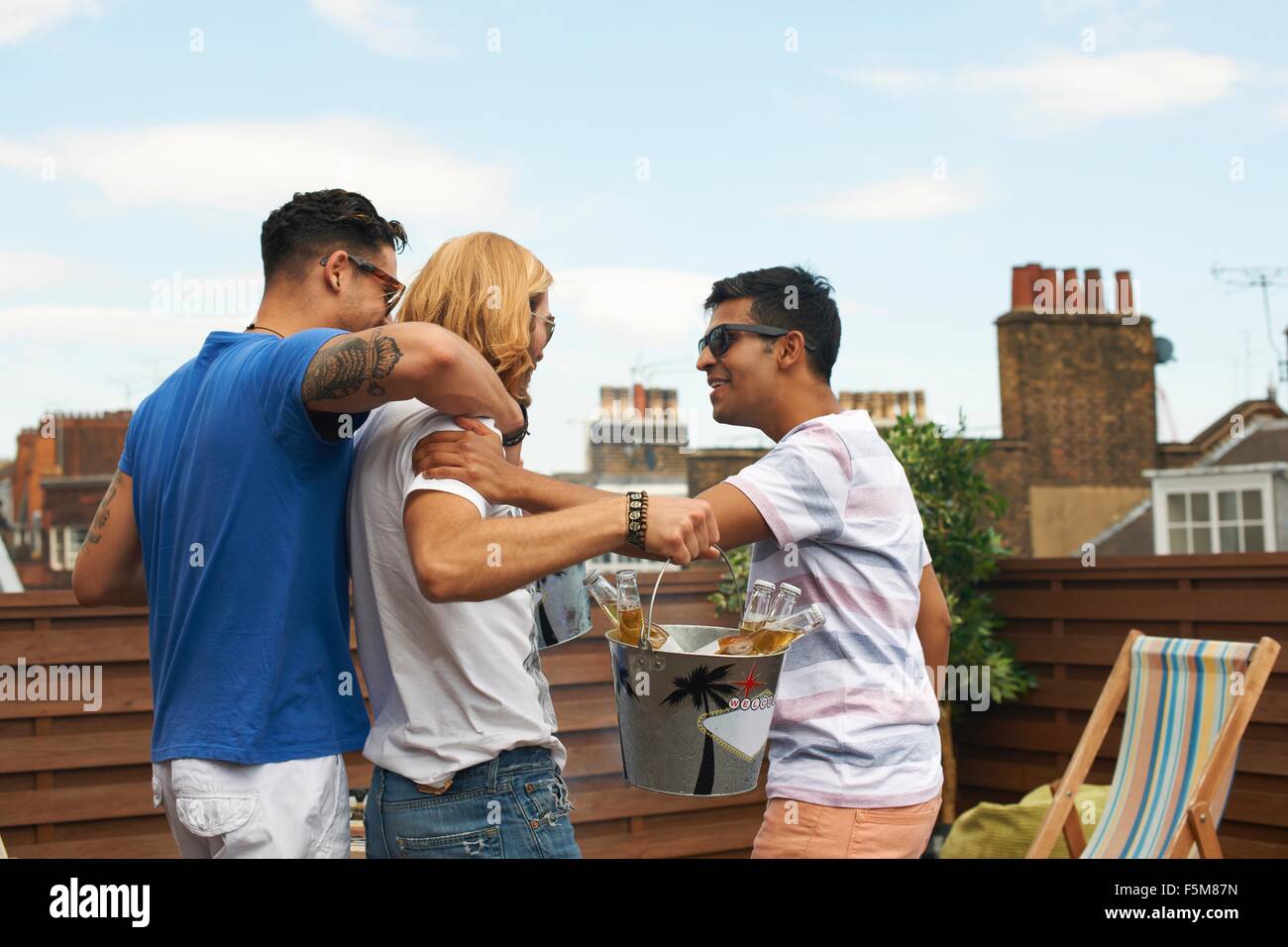 Three male friends carrying ice bucket with bottled beer at rooftop party Stock Photo