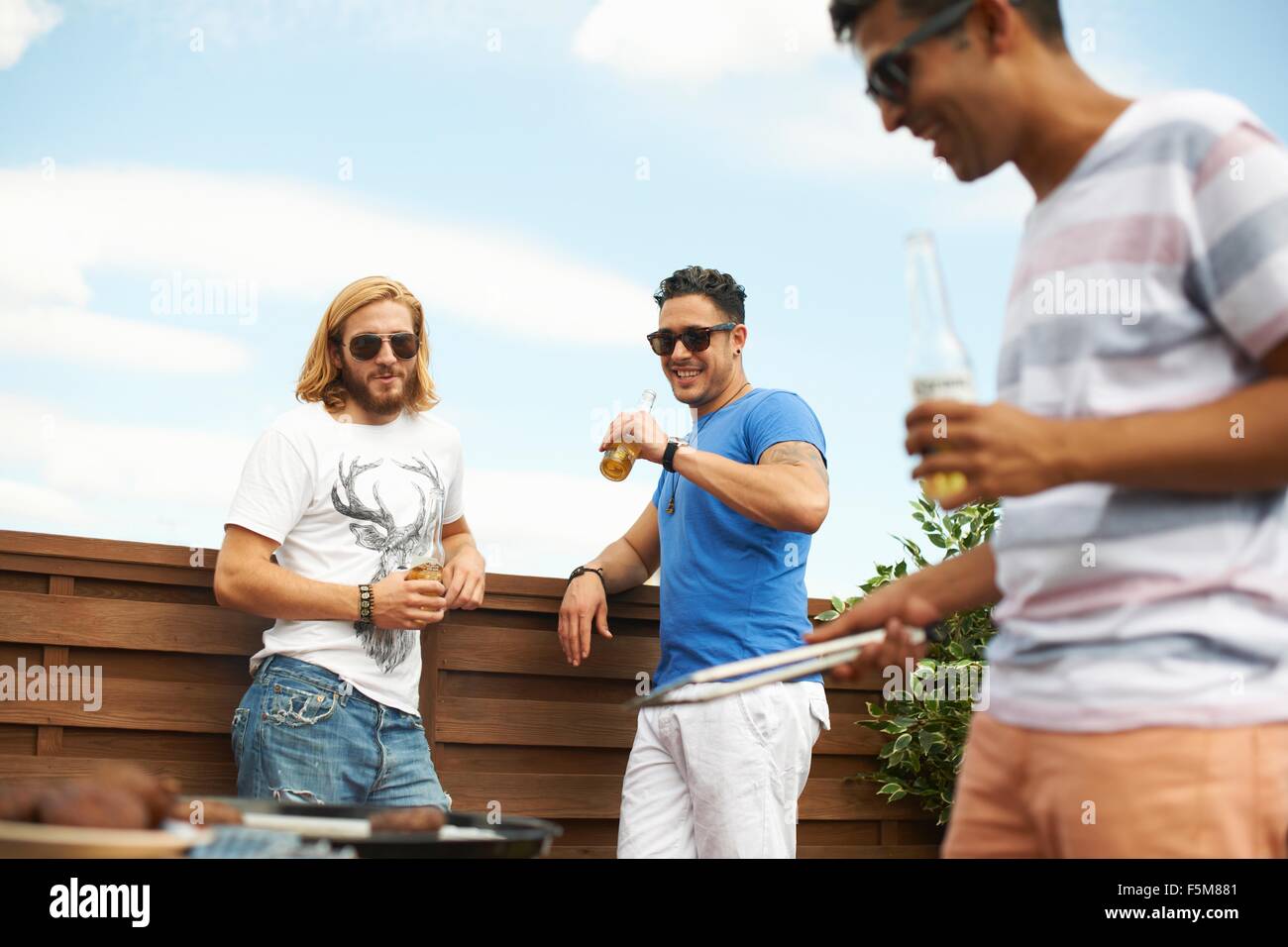 Three male friends drinking beer and barbecuing at rooftop barbecue Stock Photo