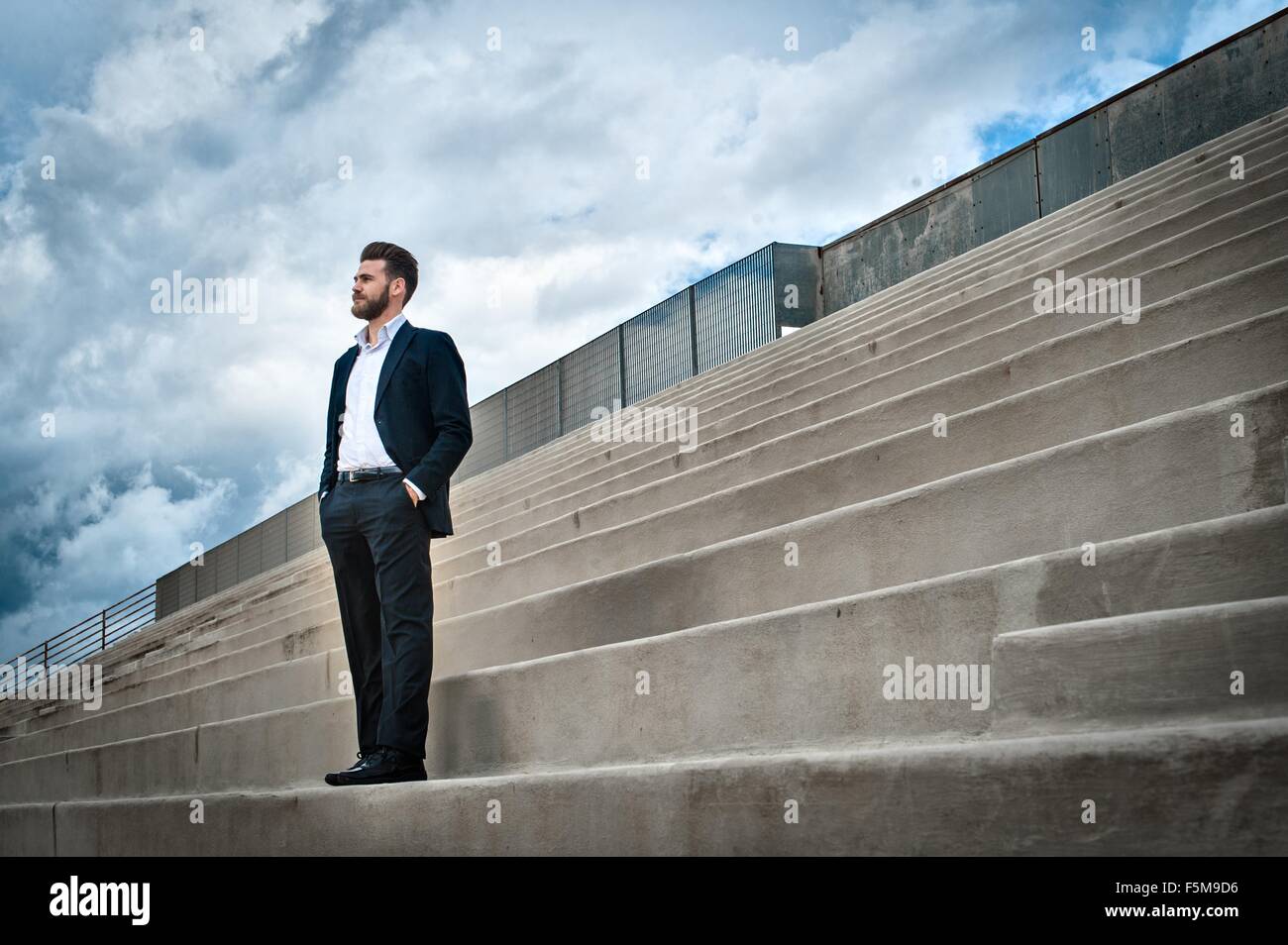 Low angle view of mid adult man on steps wearing suit, hands in pockets looking away Stock Photo