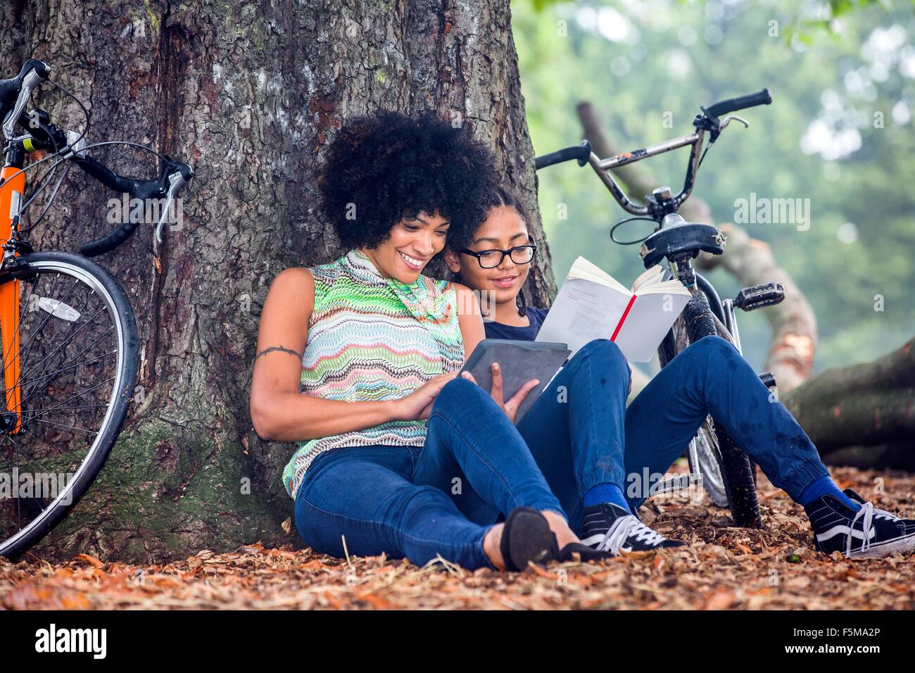 Mature woman and son sitting against park tree reading book and digital tablet Stock Photo