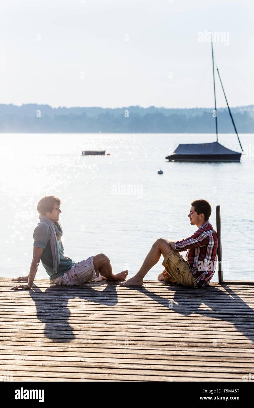 Friends sitting face to face on wood pier next to lake, Schondorf, Ammersee, Bavaria, Germany Stock Photo