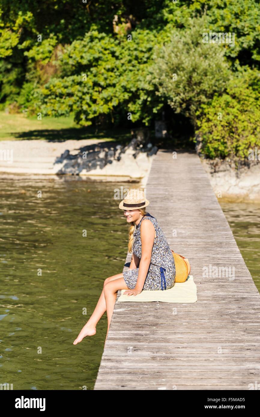Side view of young woman sitting on wood pier wearing panama hat looking away, Schondorf, Ammersee, Bavaria, Germany Stock Photo