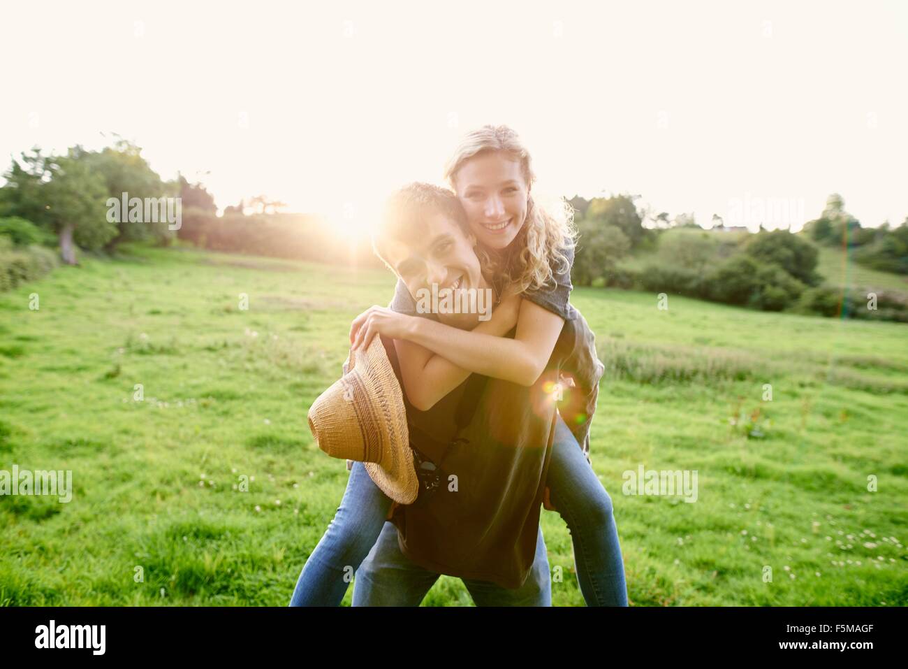 Portrait of young man giving girlfriend a piggyback in rural field Stock Photo