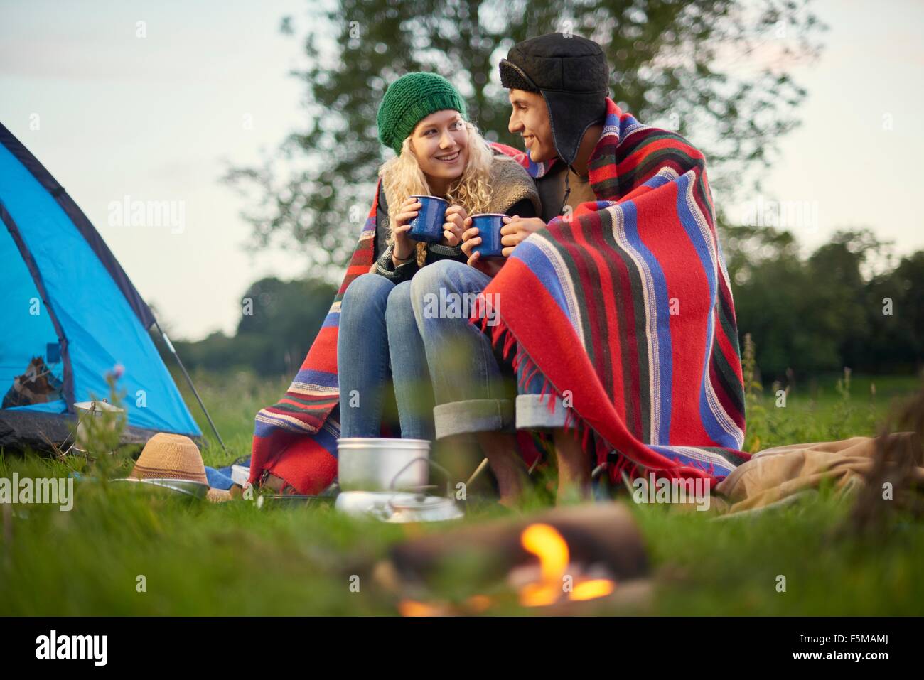 Young camping couple sitting by campfire with cup of tea Stock Photo
