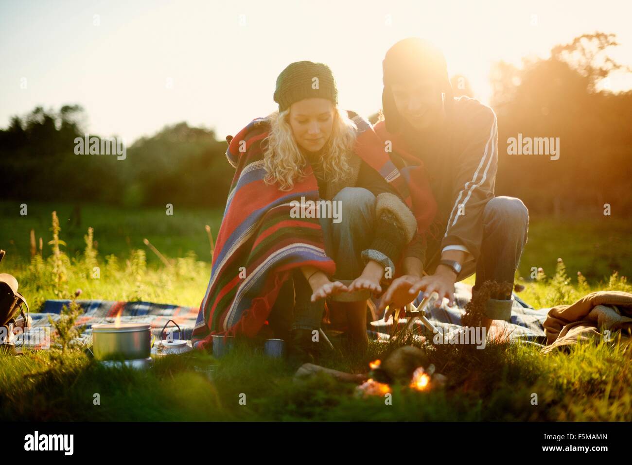Young camping couple warming hands by campfire at dusk Stock Photo