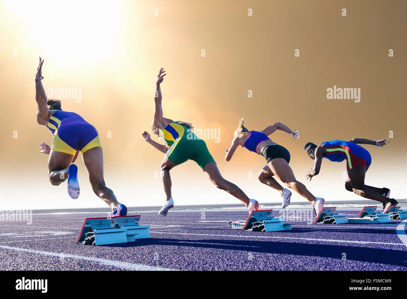 Four female athletes on athletics track, leaving starting blocks, rear view Stock Photo