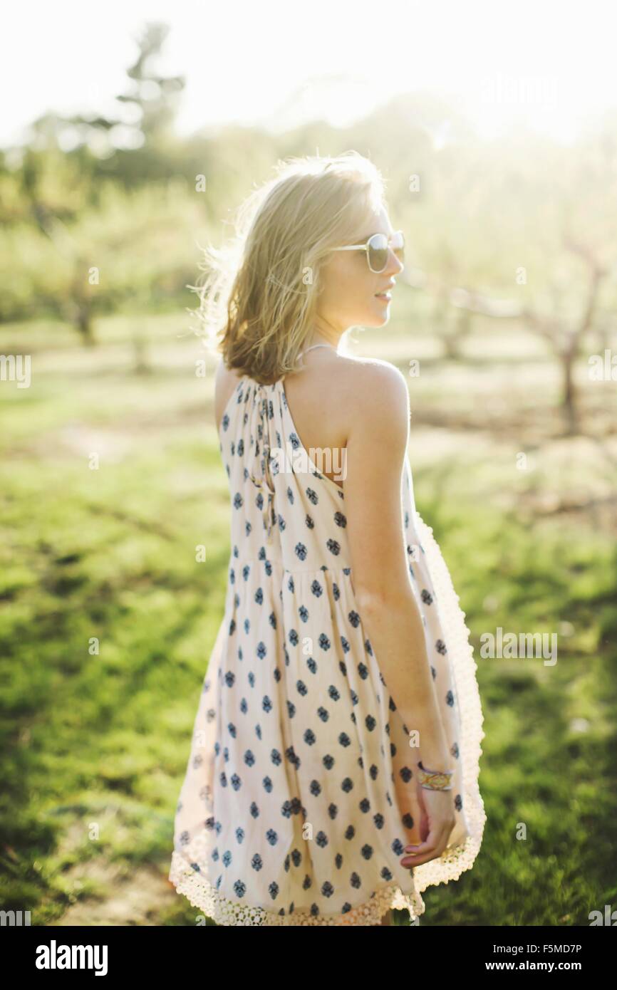 Rear view of young woman in orchard wearing sleeveless dress and sunglasses looking away Stock Photo