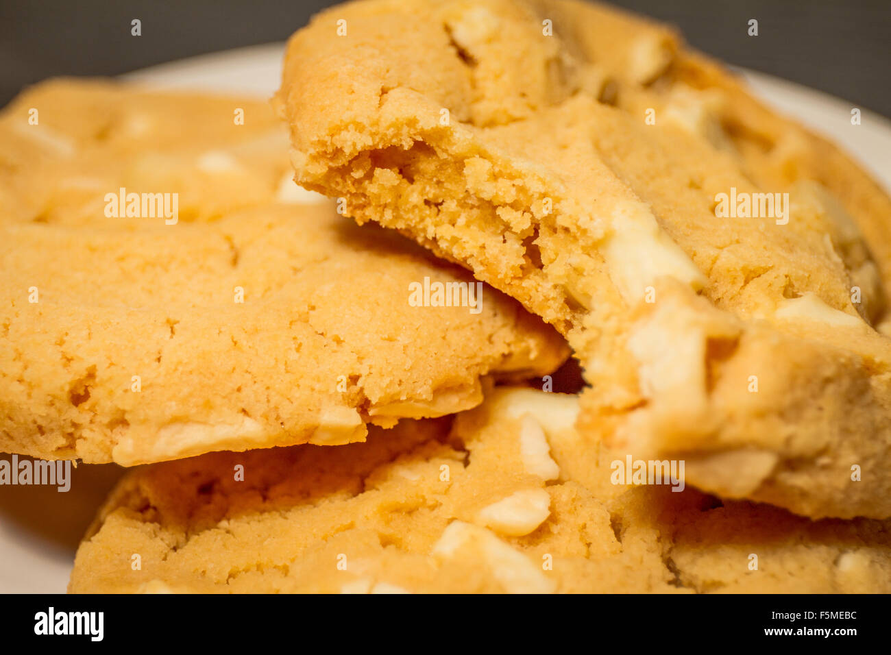 A close-up of three white chocolate chip macadamia nut cookies on a white plate with a bite taken out of one. Stock Photo