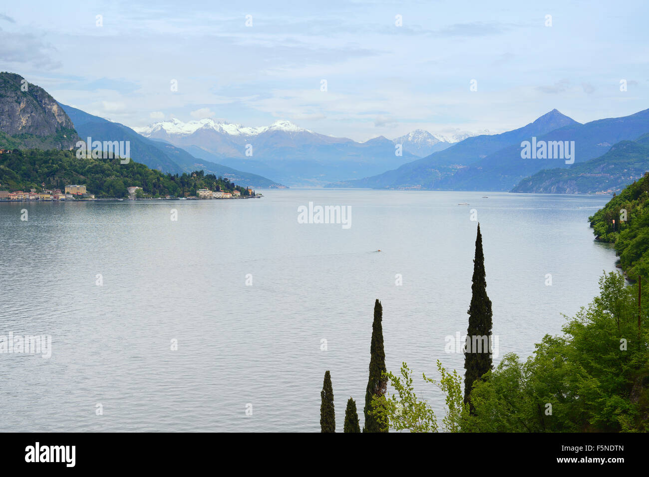 Como Lake landscape. Cypresses Trees, water, a small boat and mountains covered by snow on background. Taken near Bellagio, Ital Stock Photo