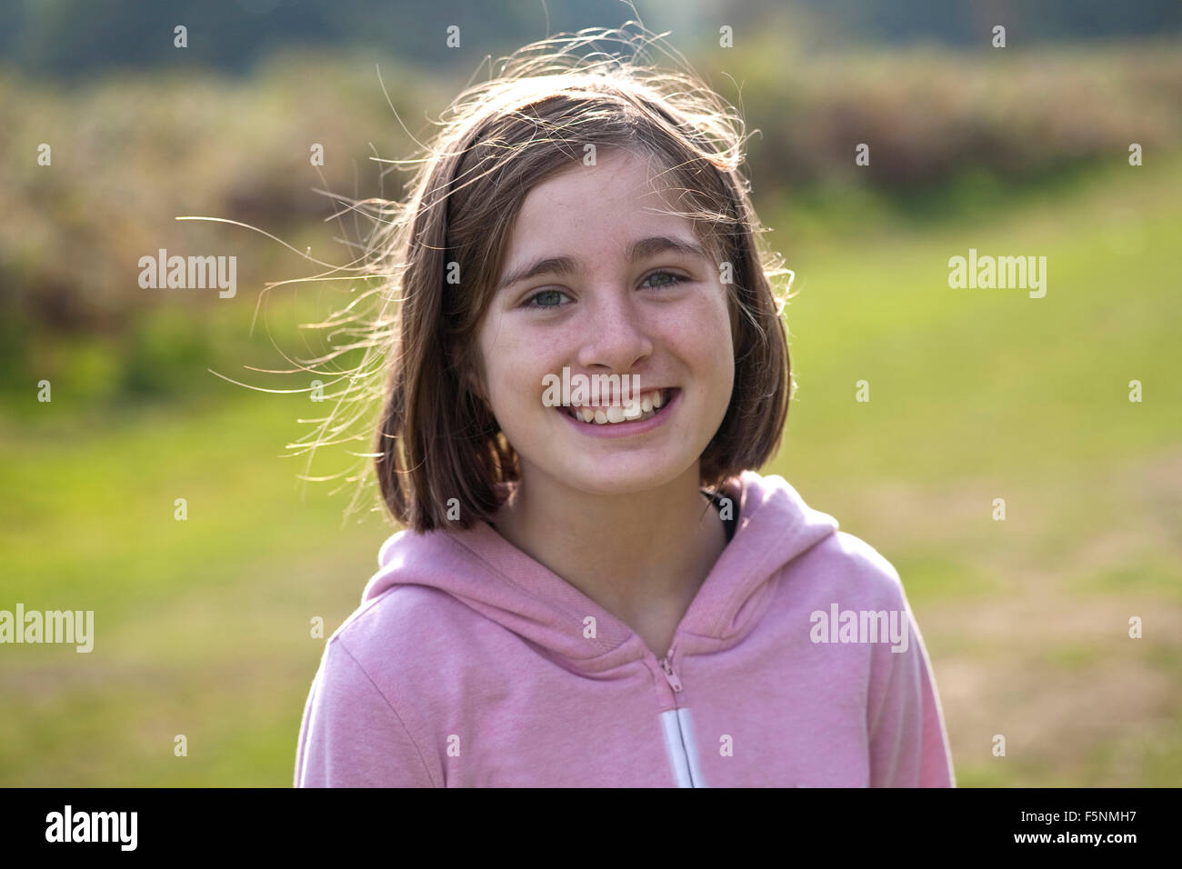A young girl smiles at the camera. She is back lit and she is standing in the middle of a park. The sunlight is autumnal. Stock Photo