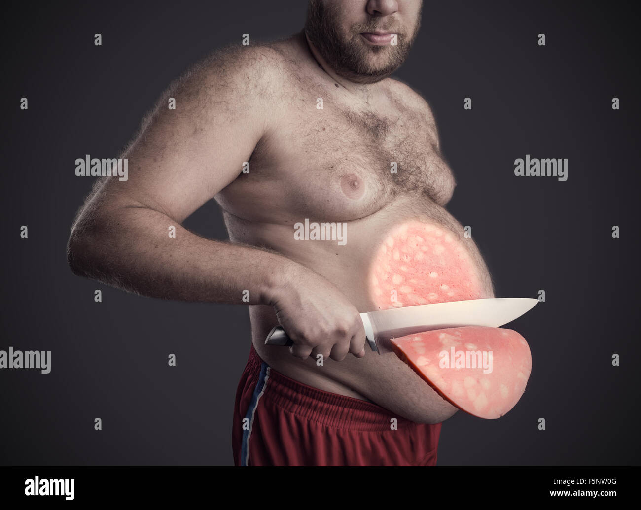 Fat man cuts a big slice of a sausage from his belly over grey Stock Photo