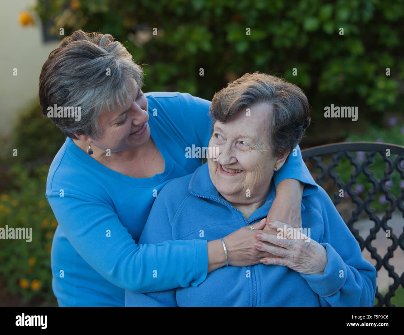 Daughter embraces mother with hands clasped who is sitting on a wrought iron bench in the garden. Stock Photo