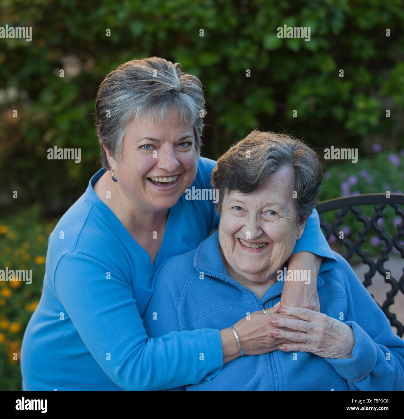 Daughter and Mother Laughing and embracing in the Garden with mother sitting on wrought iron bench and daughter's arms are around her. Stock Photo