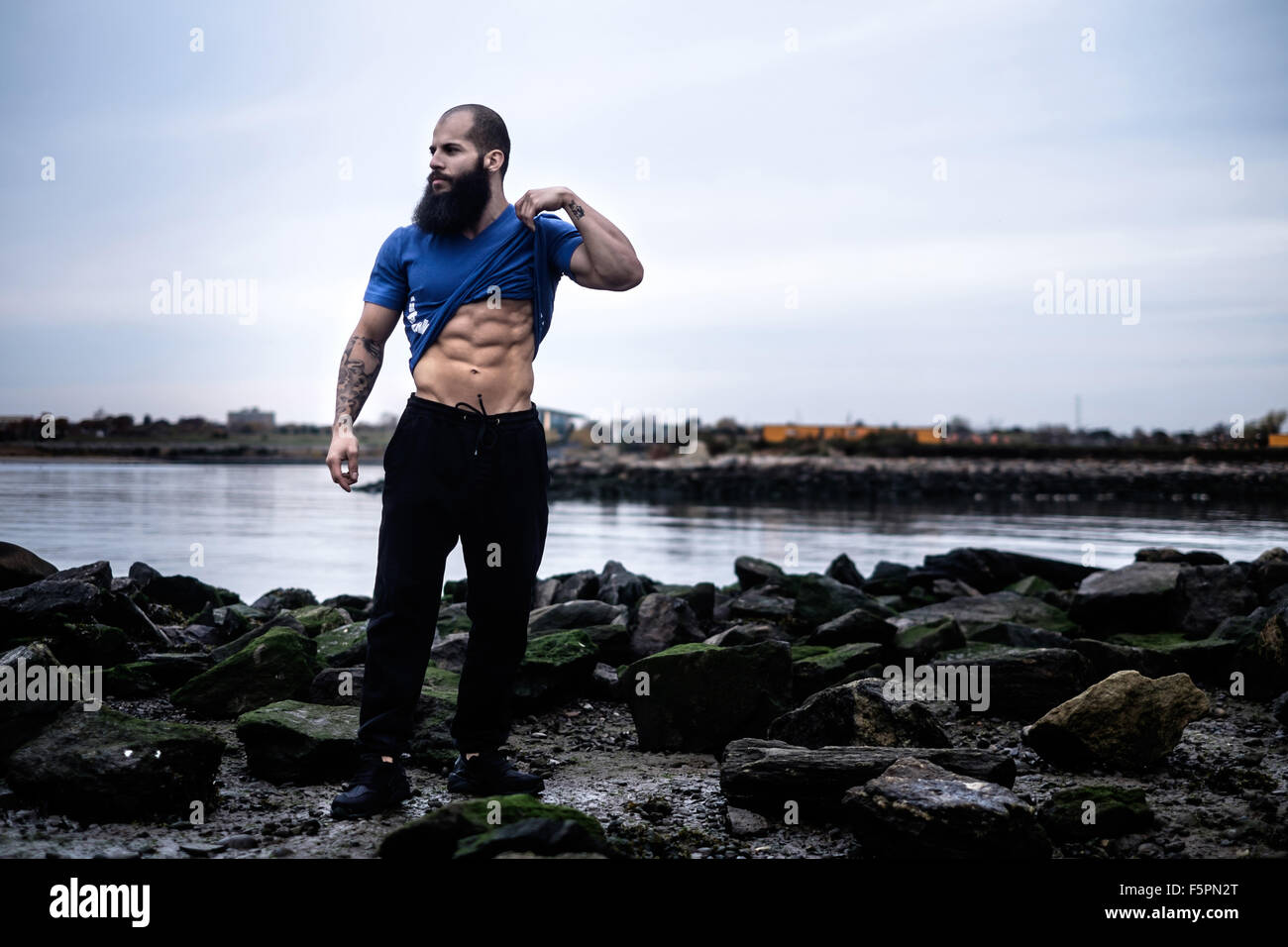 Man with a beard lifts his shirt to reveal abdominal muscles on a rocky beach. Stock Photo