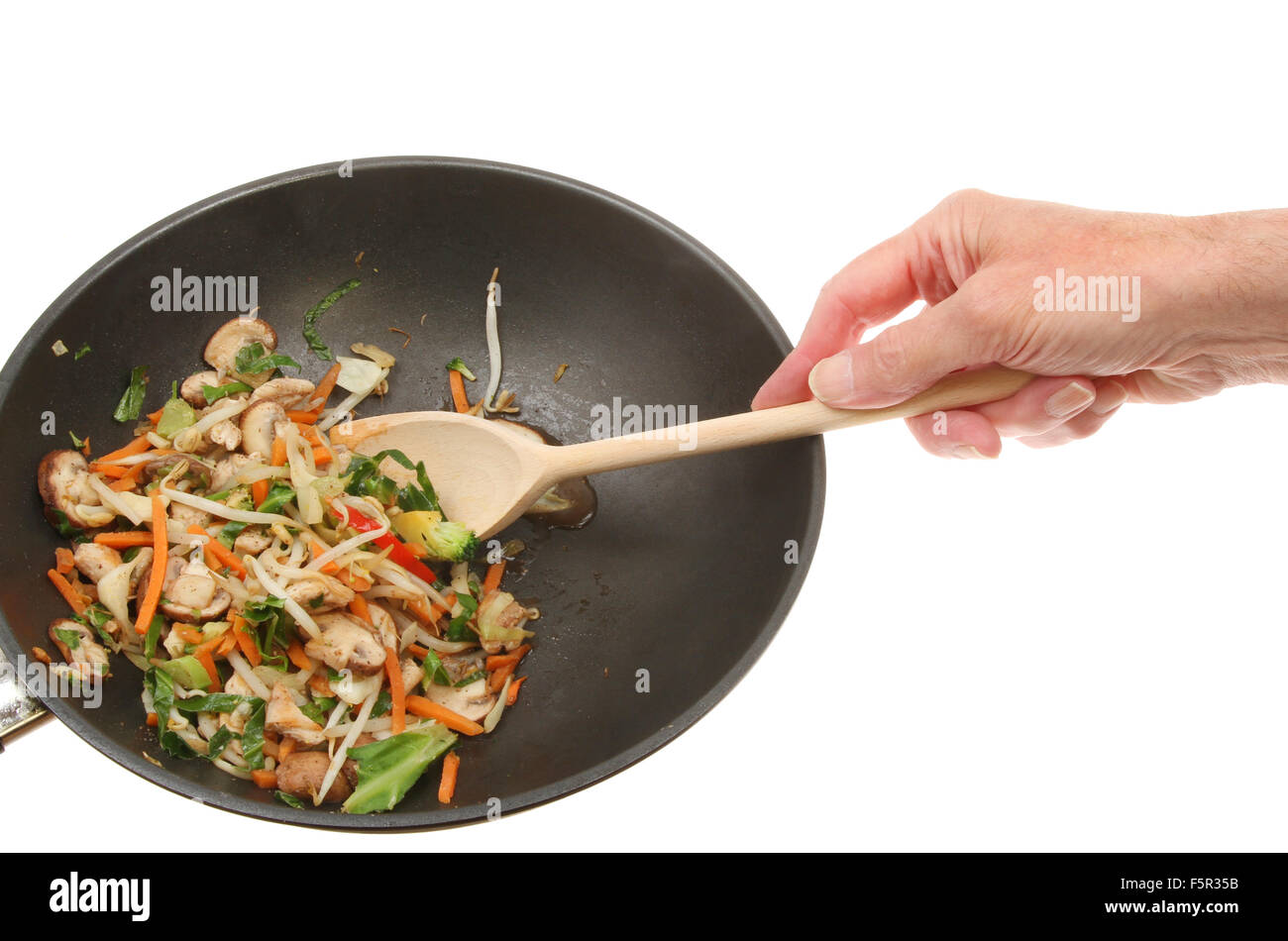 Hand with wooden spoon stirring chicken and stir fry vegetables in a wok against a white background Stock Photo