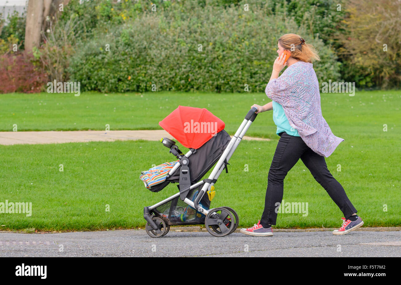 Young woman pushing a pushchair through a park while speaking on a mobile phone. Woman walking with baby using smartphone. Stock Photo