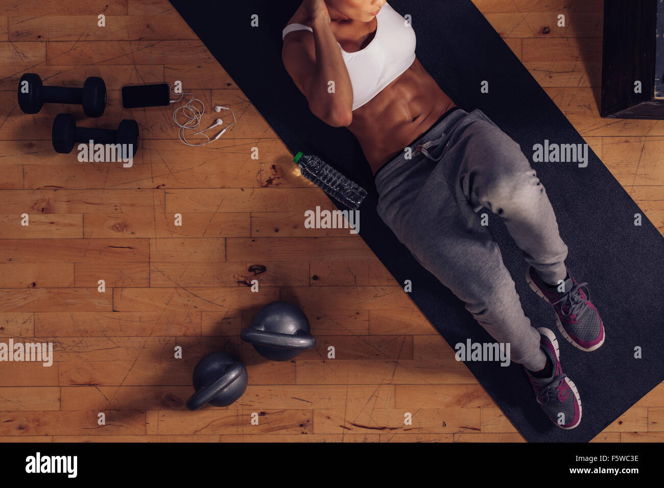 Top view of muscular woman doing sit-ups. Female lying on exercise mat doing stomach workout with gym equipments in floor. Stock Photo
