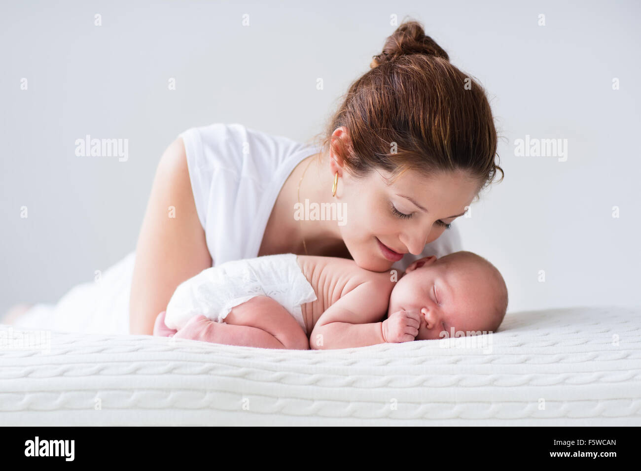 Young mother hugging her newborn child. Mom nursing baby. Woman and new born boy relax in a white bedroom. Family at home. Stock Photo