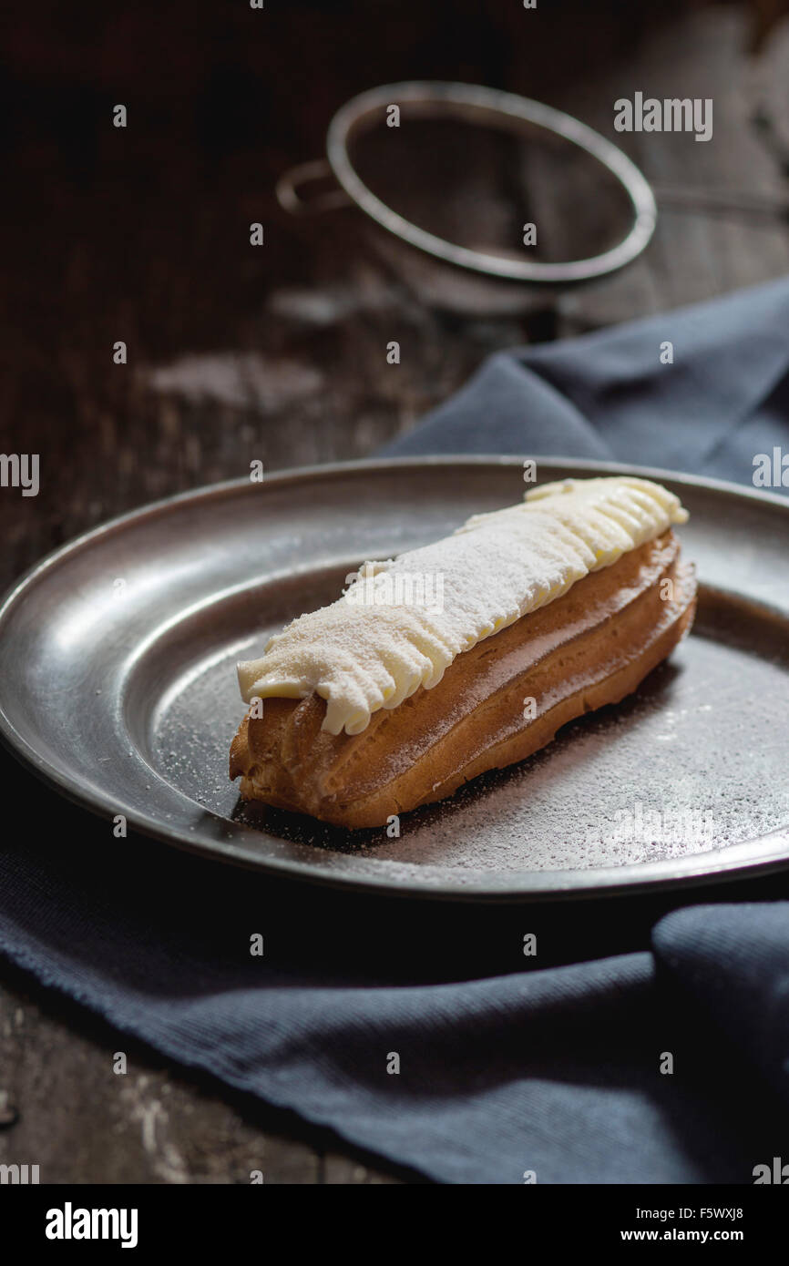 Single Eclair with white butter cream in vintage metal plate. Dark rustic style. Day light. Outside view Stock Photo