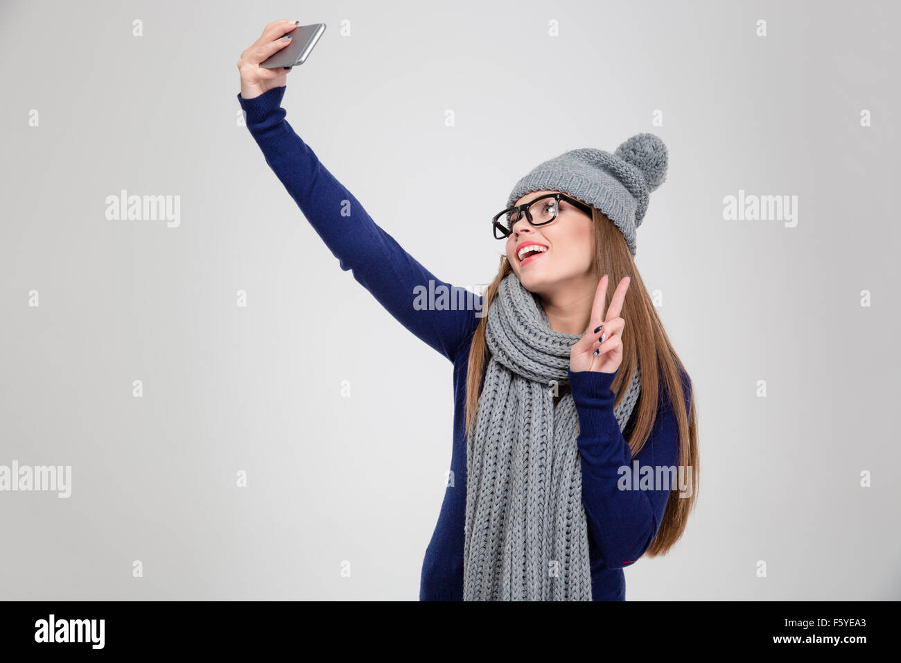 Portrait of a happy woman in winter cloth making selfie photo on smartphone and showing peace sign isolated on a white backgroun Stock Photo