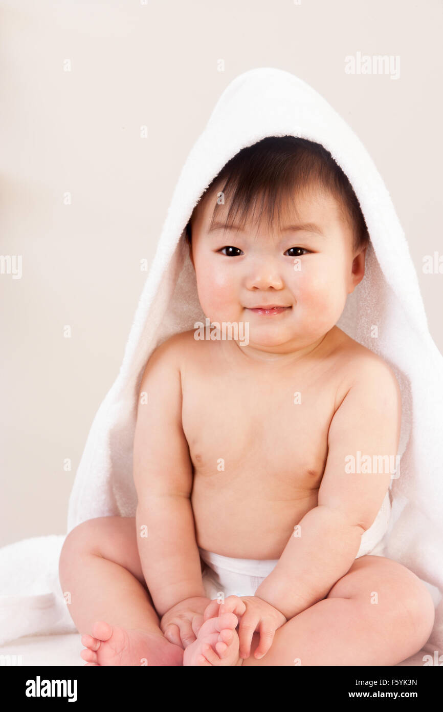 Cute Asian baby sitting with a towel over his head. He is wearing a nappy and is smiling at the camera. Stock Photo