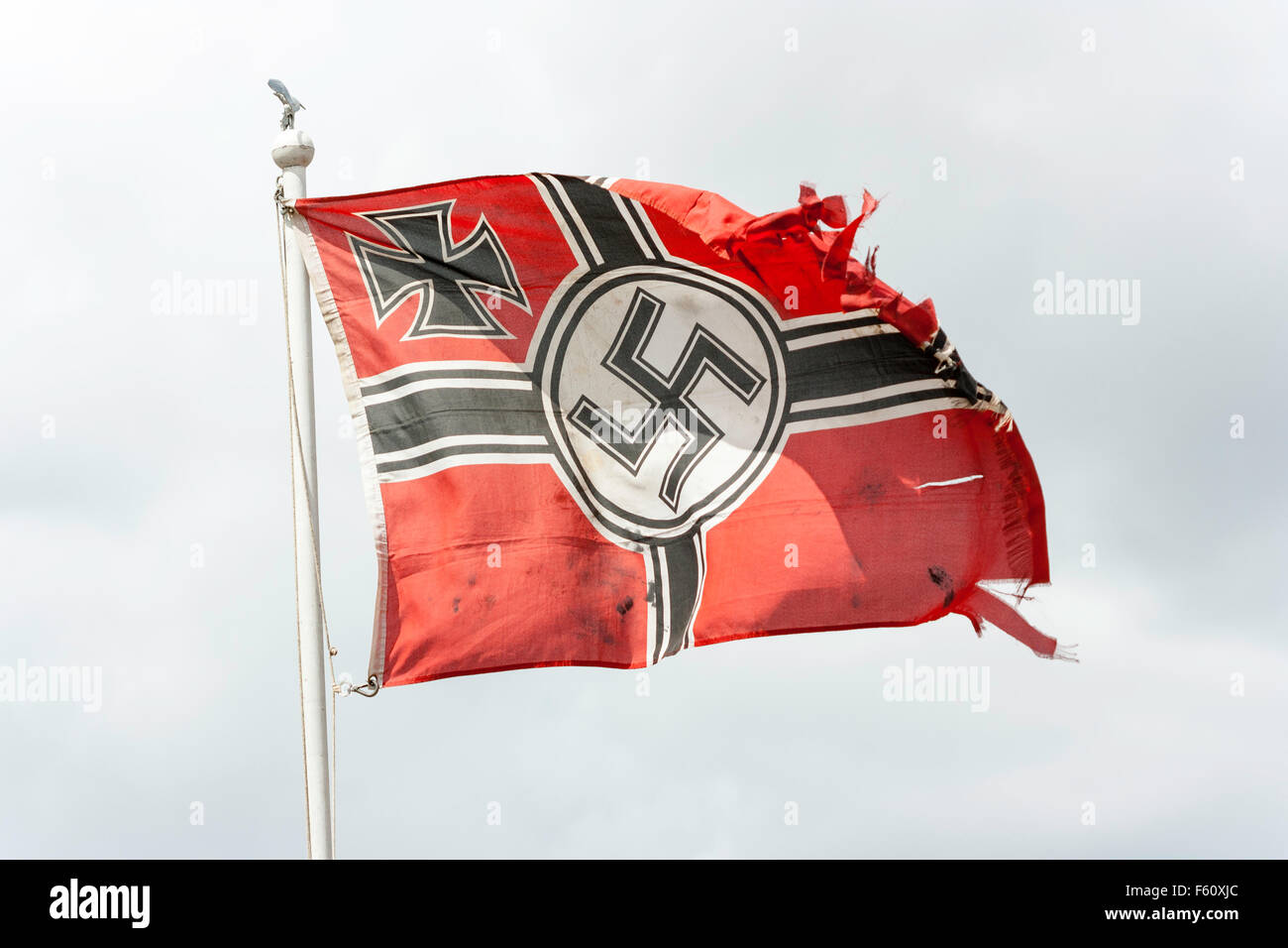 World war two Re-enactment. War torn German Wehrmacht flag with Iron cross and Nazi swastika, fluttering on top of flagpole against stormy grey sky. Stock Photo