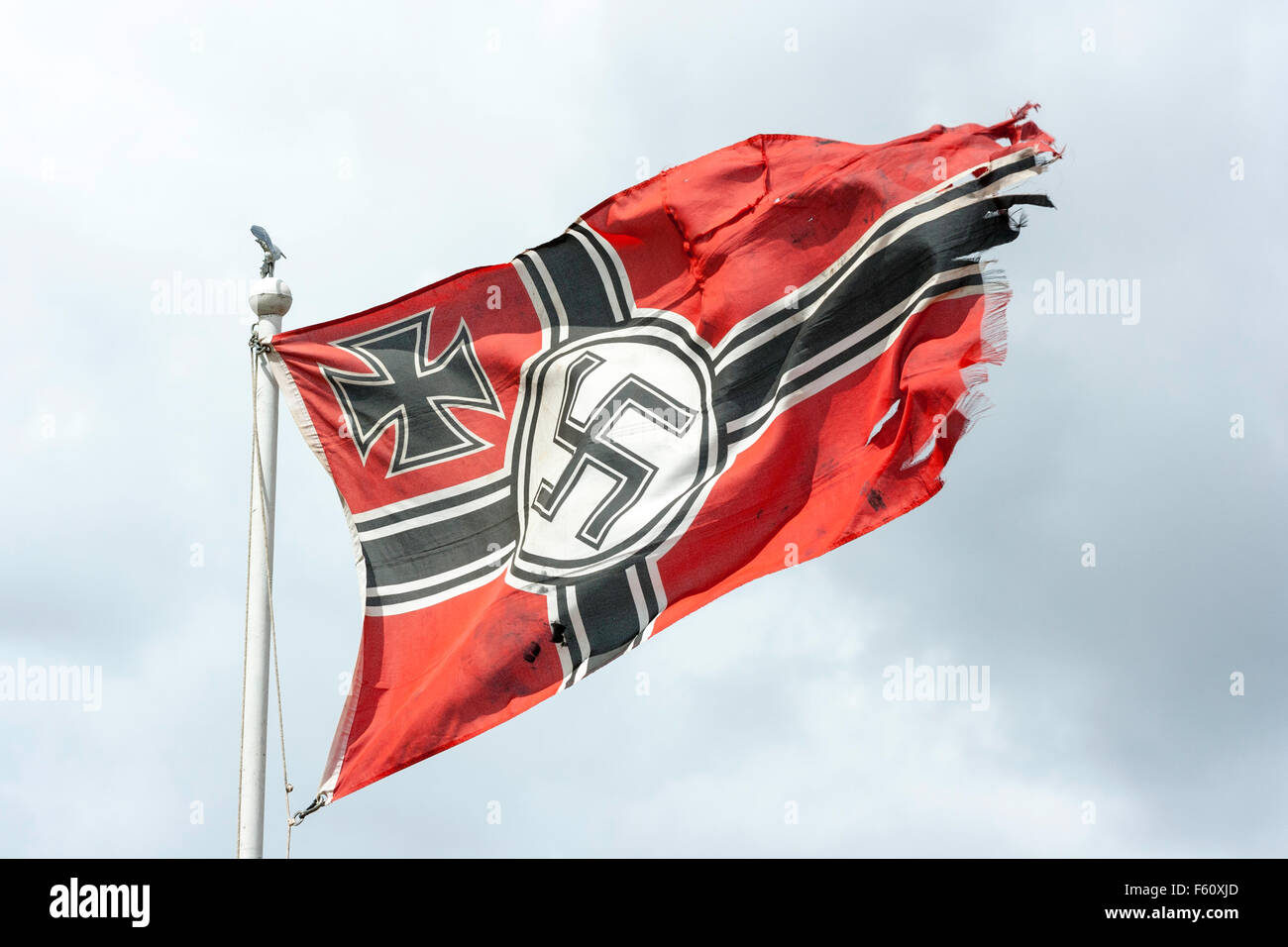 World war two Re-enactment. War torn German Wehrmacht flag with Iron cross and Nazi swastika, fluttering on top of flagpole against stormy grey sky. Stock Photo