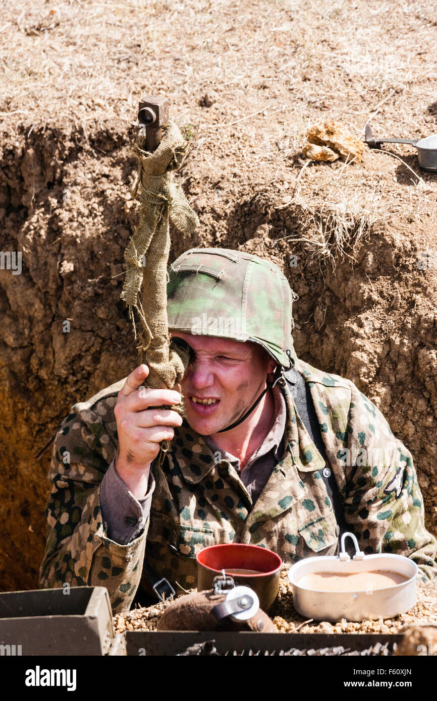 World war two re-enactment. View facing looking down at German officer wearing camouflage smock, standing in trench looking through periscope. Stock Photo