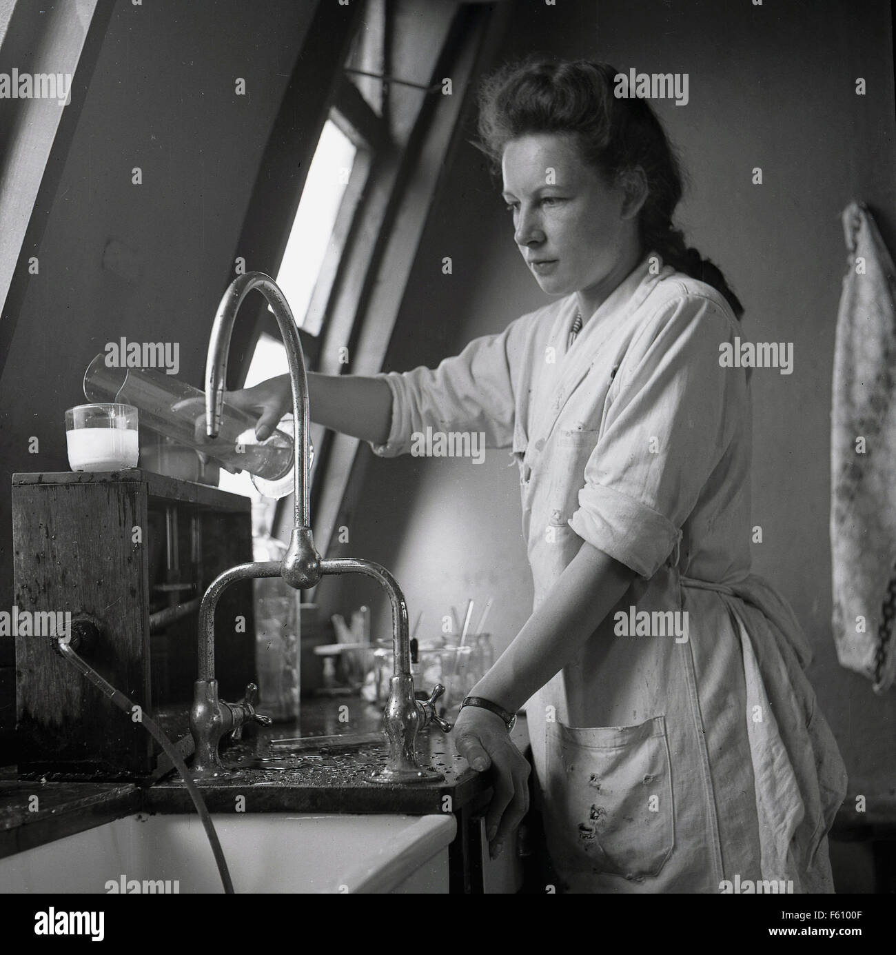 1950s historical picture, white coated young female adult student doing a chemical experiment in a small top floor laboratory. Stock Photo