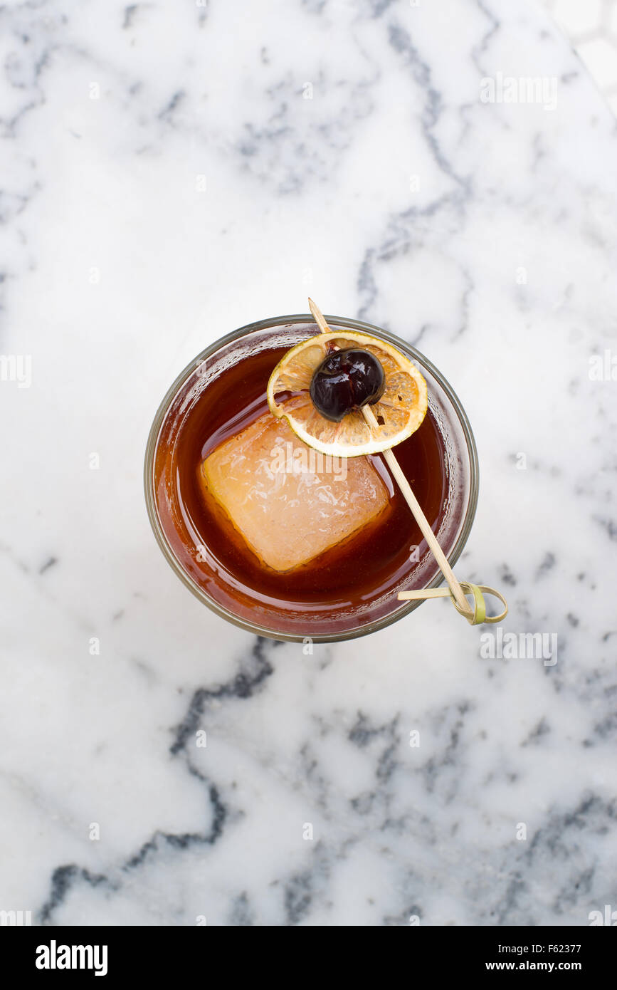 Overhead view of a coffee-colored cocktail with dark cherry and lime garnish on a white marble table top. Stock Photo