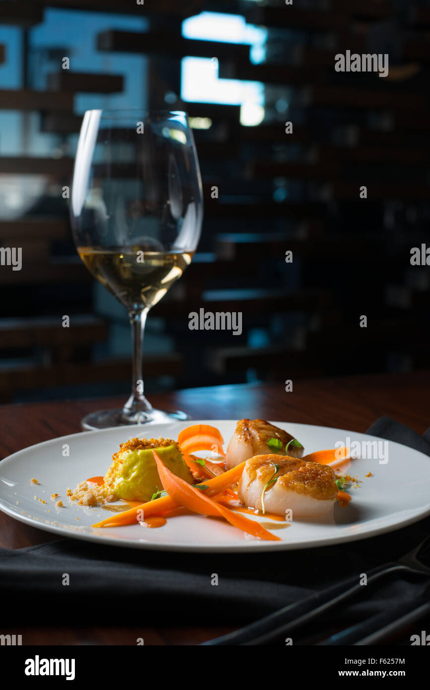 Scallops with yellow mousse and a glass of white wine in the background in a dark restaurant setting. Stock Photo