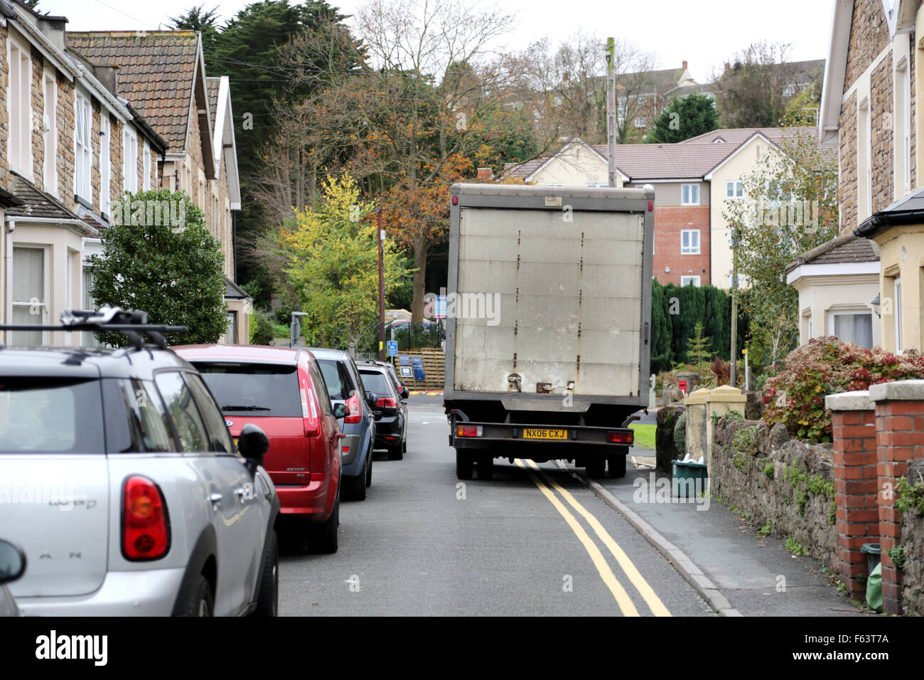 A lorry is forced to drive on the pavement or mount the kerb to pass parked cars on a narrow British street. The road is in a residential area Stock Photo