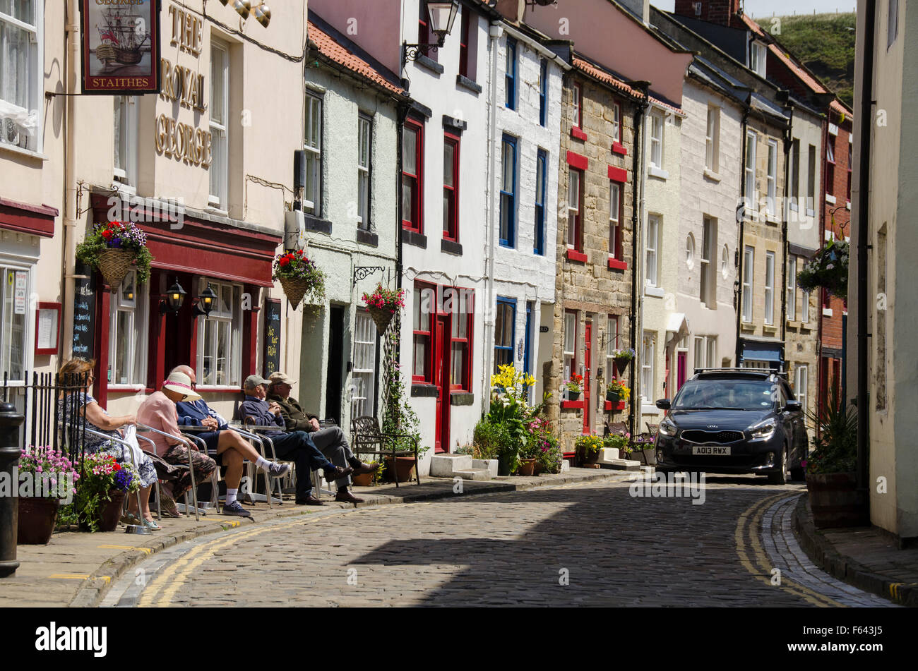 Car rounds the corner as people sit in the sun, relaxing outside pub - narrow cobbled High Street, quaint village of Staithes, North Yorkshire, UK. Stock Photo