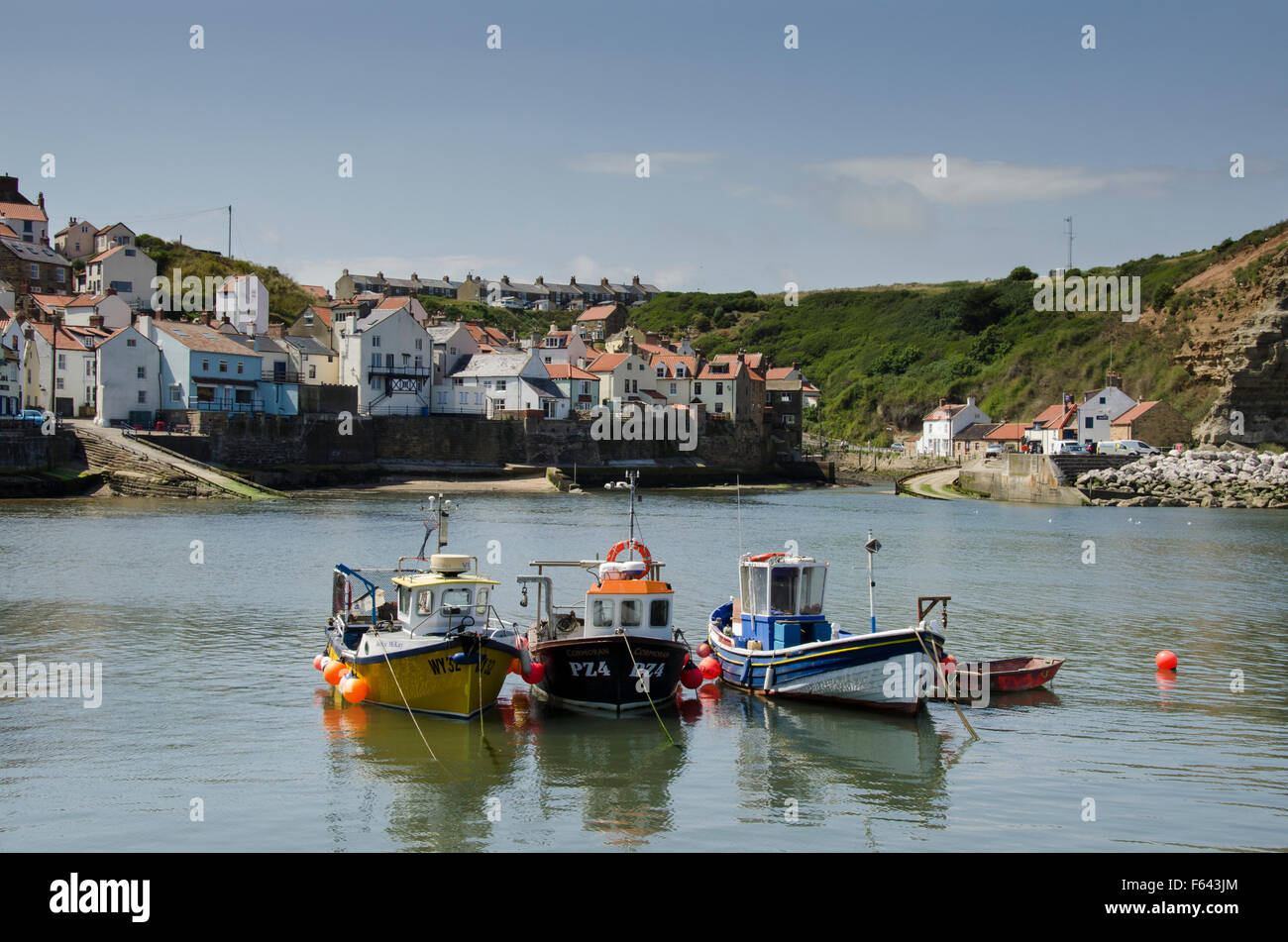 Sunny summer day with blue sky, 3 colourful fishing boats moored in the harbour with the picturesque village of Staithes, North Yorkshire, UK, beyond. Stock Photo