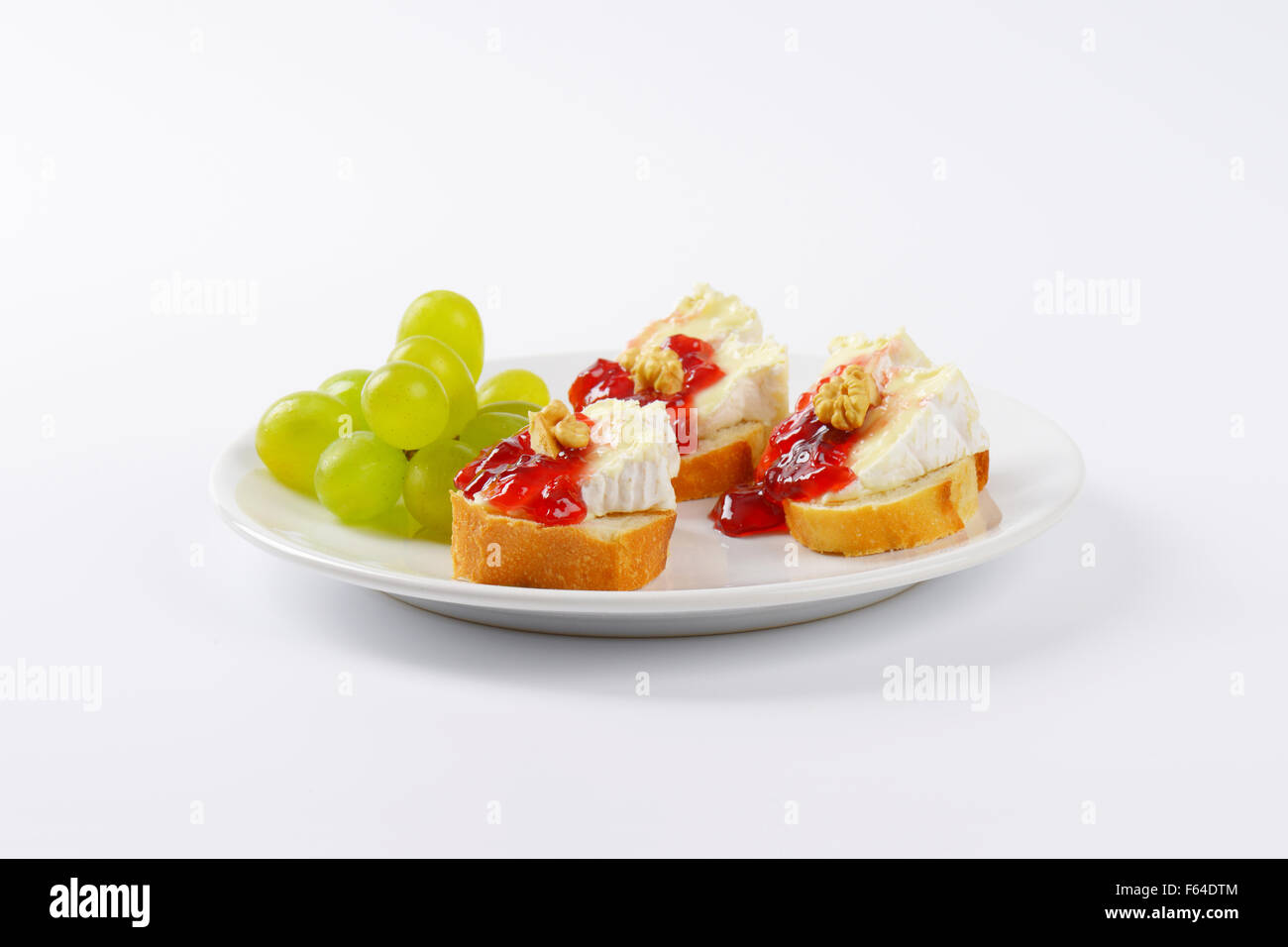 slices of fresh bread with brie cheese, walnuts, jam and grapes on white plate Stock Photo