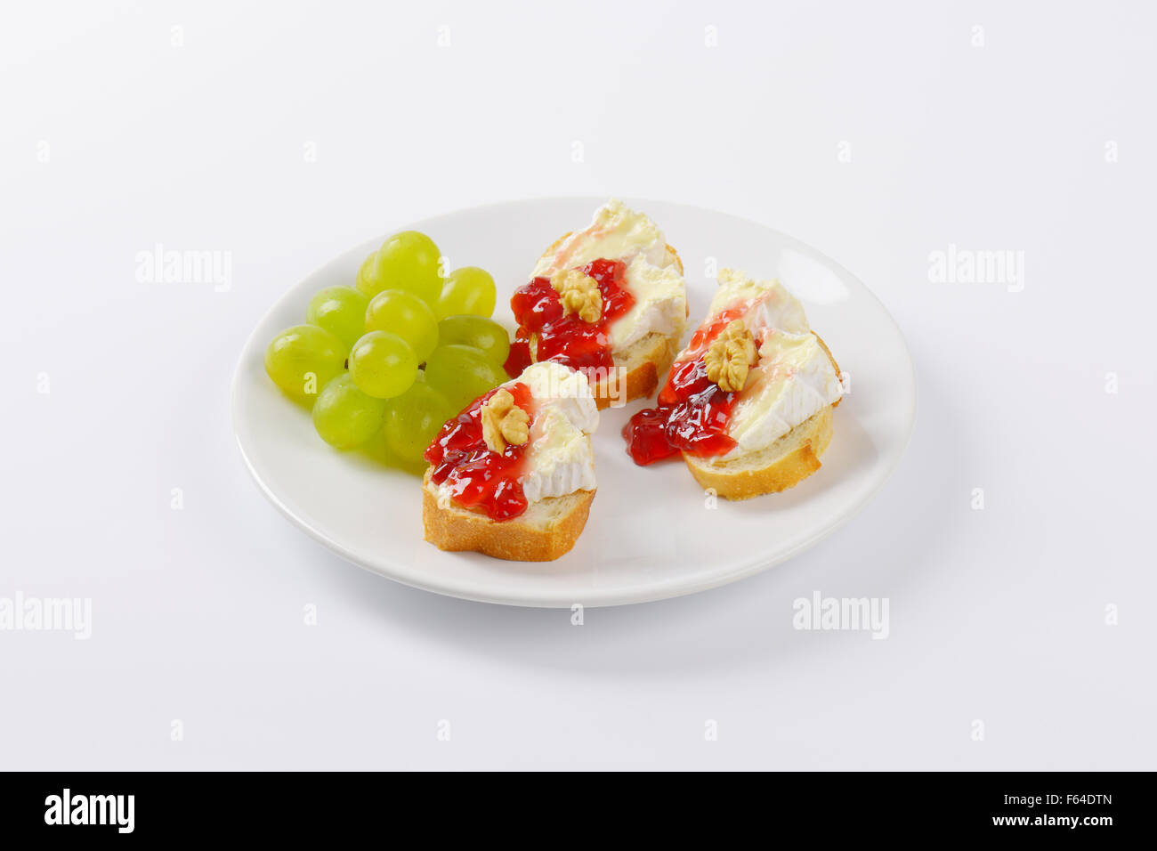 slices of fresh bread with brie cheese, walnuts, jam and grapes on white plate Stock Photo